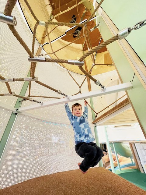A young boy hanging from a horizontal bar in an indoor play area with ropes and climbing structures.