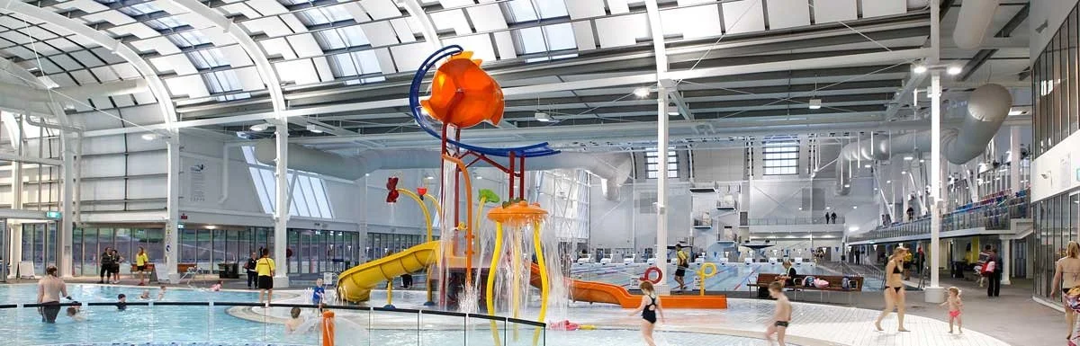 Indoor water park featuring a large splash pad with slides, children playing, and visitors relaxing.
