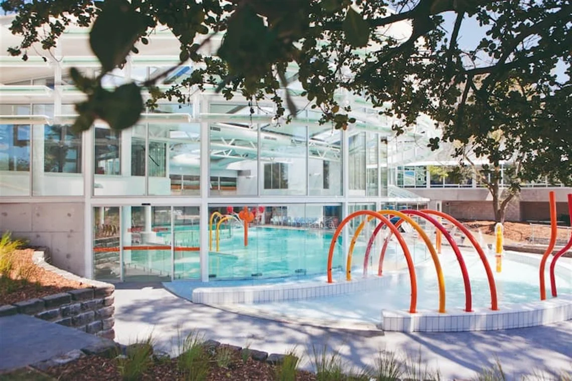 Outdoor area of a modern building with a swimming pool, glass windows, and orange and yellow water spray features, shaded by trees.