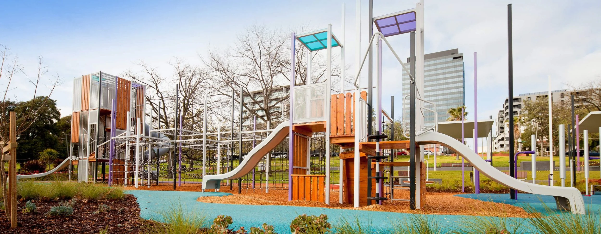 Colorful playground with slides, climbing structures, and various play equipment, surrounded by greenery and trees in an urban park setting.