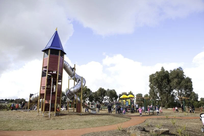 Children playing on a large playground with a blue-roofed tower and slides, surrounded by trees and people.