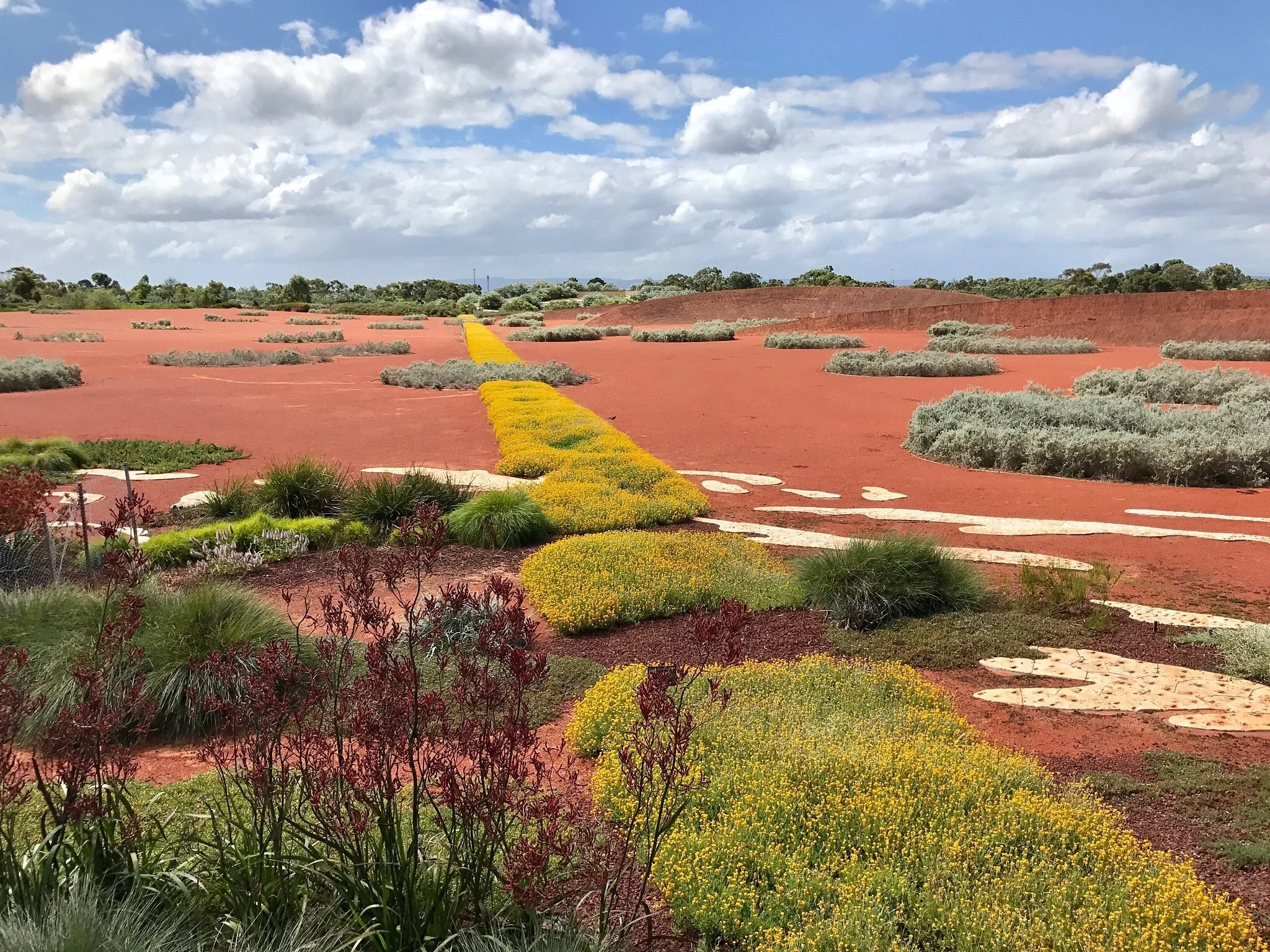 A garden with colorful plants along a curved pathway in a vast red desert landscape under a partly cloudy sky.