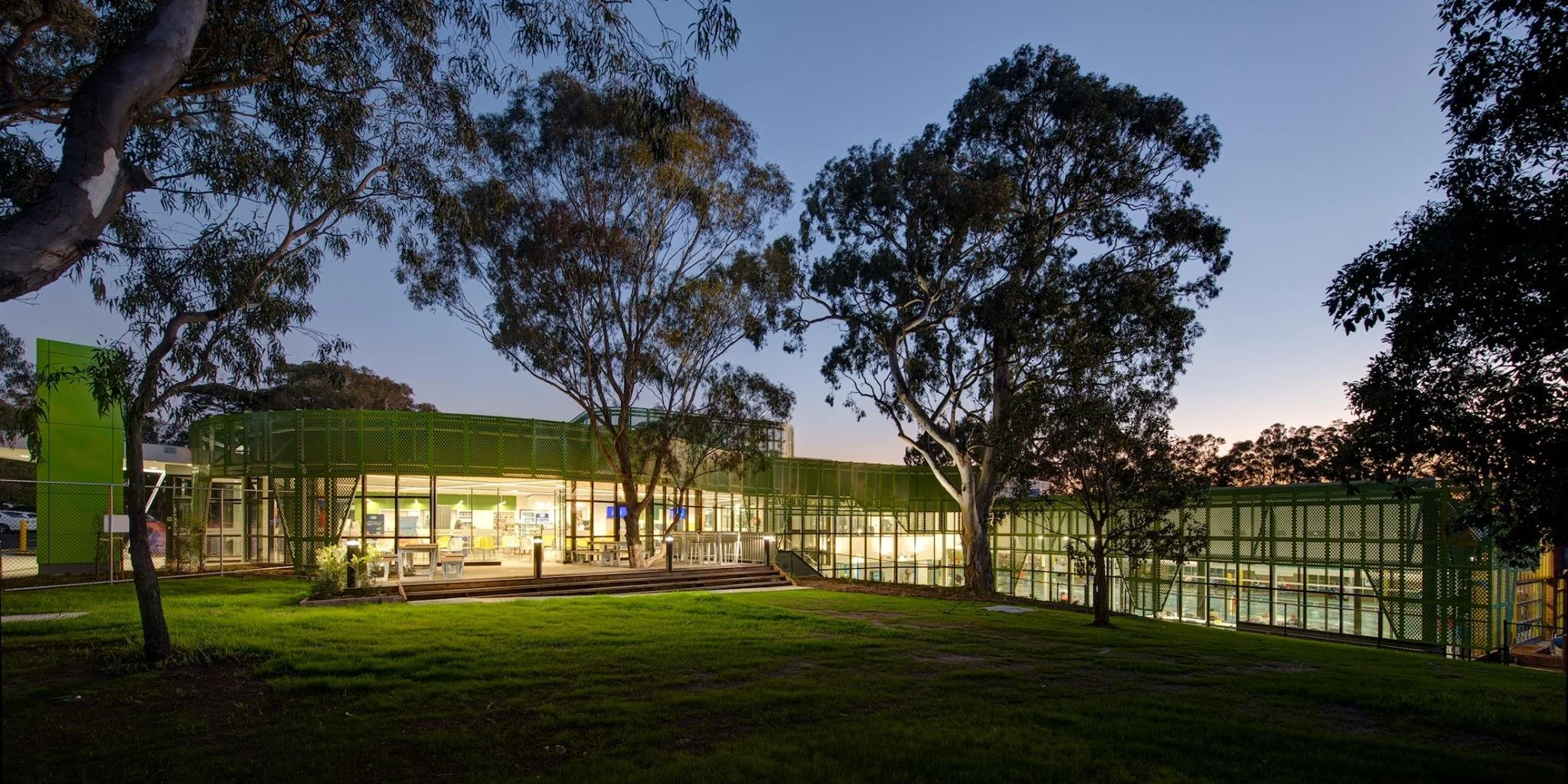 A modern building with green metal accents is illuminated at dusk, surrounded by trees and a grassy area.