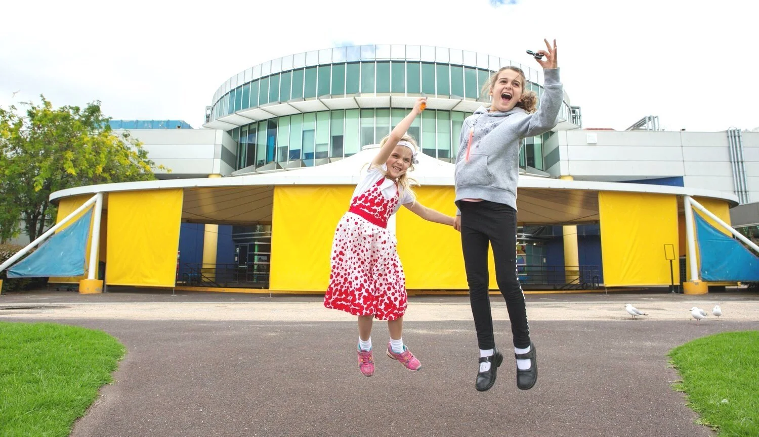 Two girls jumping and smiling in front of a modern building with yellow and blue accents, holding hands on a paved walkway surrounded by green grass.