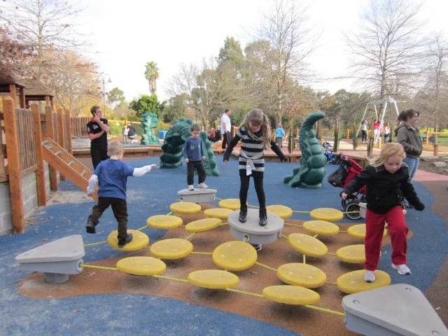Children playing on a playground with stepping stones and climbing features, surrounded by trees and adults supervising.