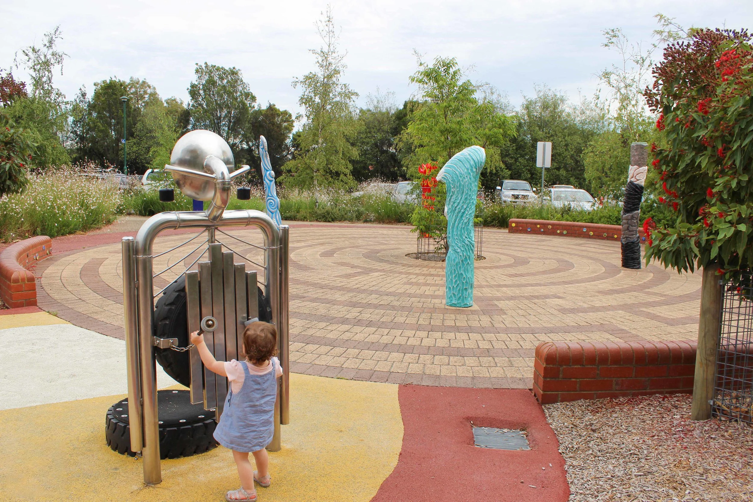 A playground with sculptures and a girl playing with a tire swing, surrounded by trees and parking lot.