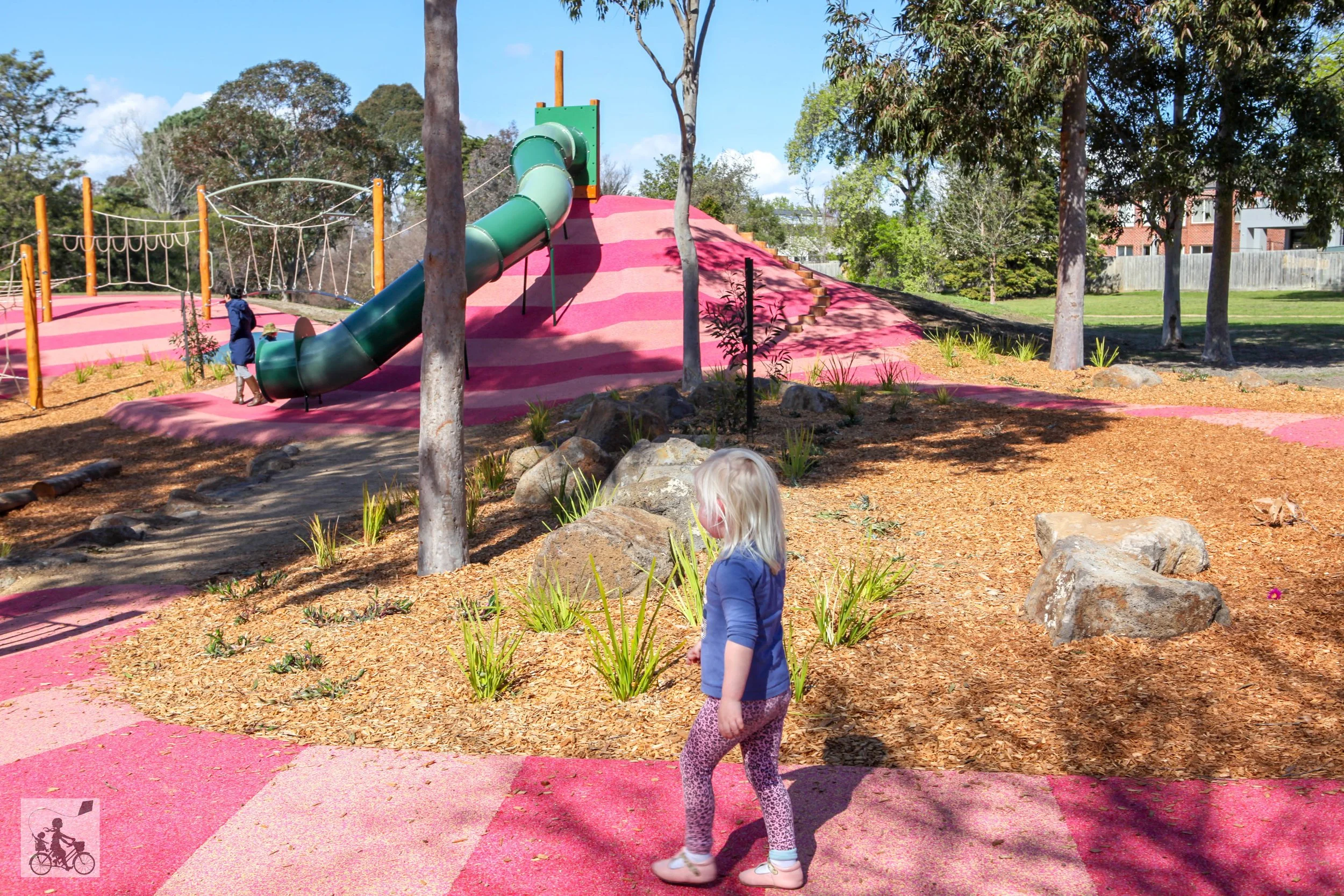 A playground with a pink and purple slide and climbing area, surrounded by trees and rocks. Two children are playing, one with a dog and the other observing.