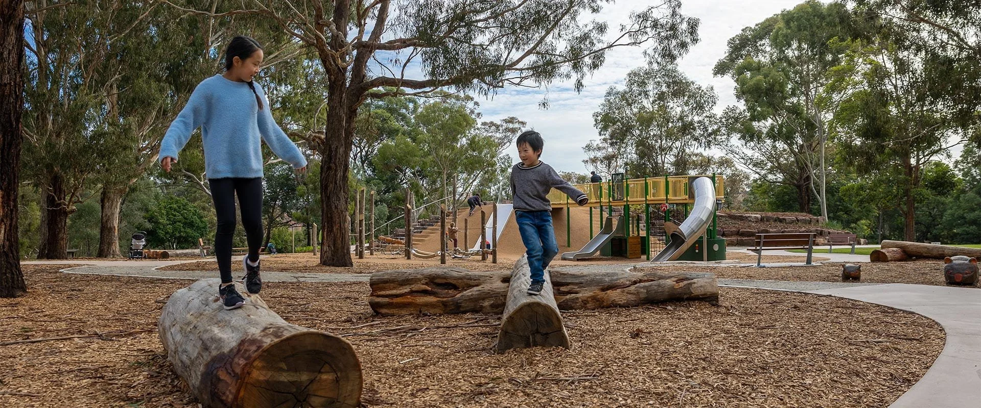 Two children balancing on logs in a park playground with trees and a slide in the background.