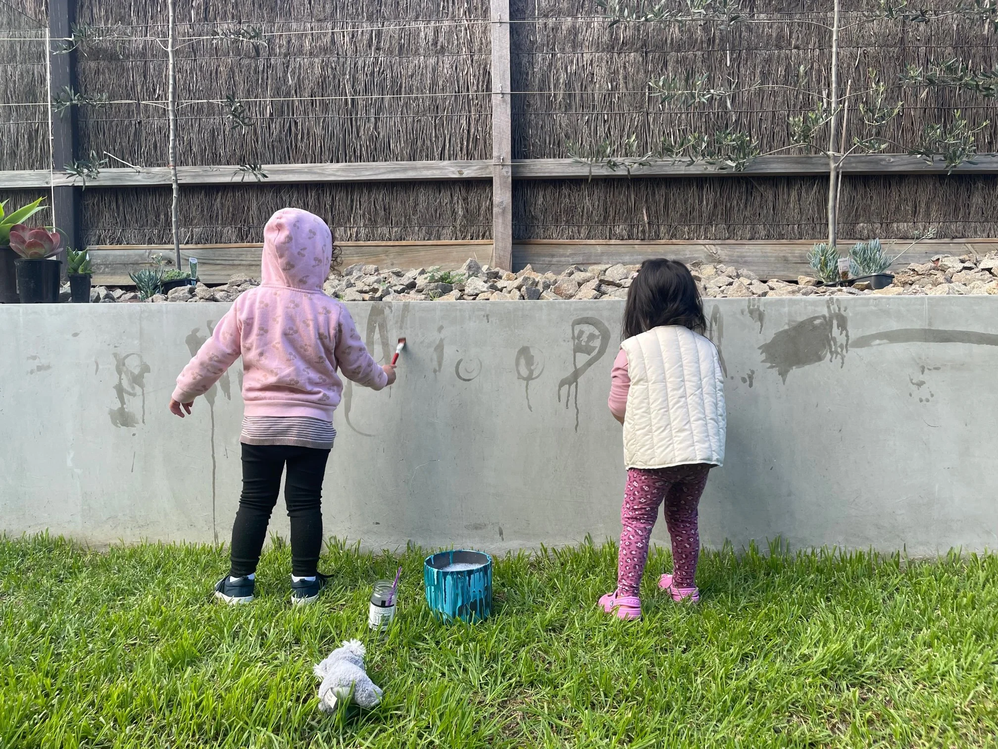 Two young girls with dark hair, one wearing a pink hoodie and black pants, the other wearing a white quilted vest and pink leggings with purple pattern, are painting or drawing on a concrete wall using brushes. There is a small bucket of paint and a stuffed animal on the grass in front of them. The background features a wooden fence with plants and small trees.