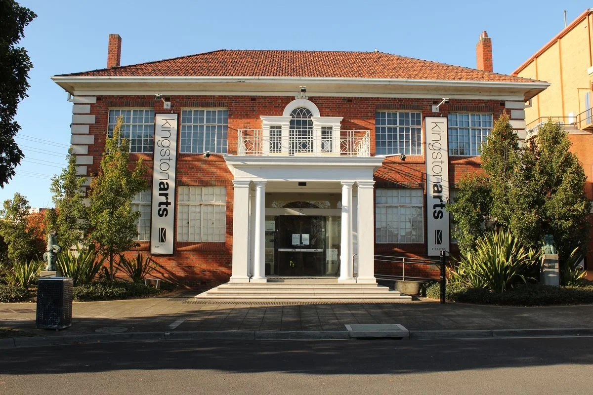 The image shows a two-story brick building with white architectural details, a red-tiled roof, and a small balcony above the entrance. There are two vertical banners on either side of the entrance that read "Kingston Arts". The building is surrounded by trees and plants, with a sidewalk and street in the foreground.