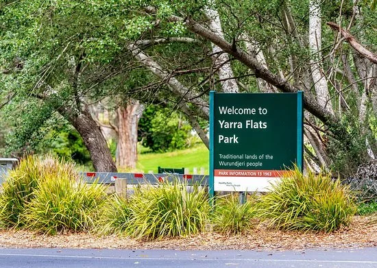 Green park sign reading 'Welcome to Yarra Flats Park, Traditional lands of the Wurundjeri People' surrounded by trees and bushes.