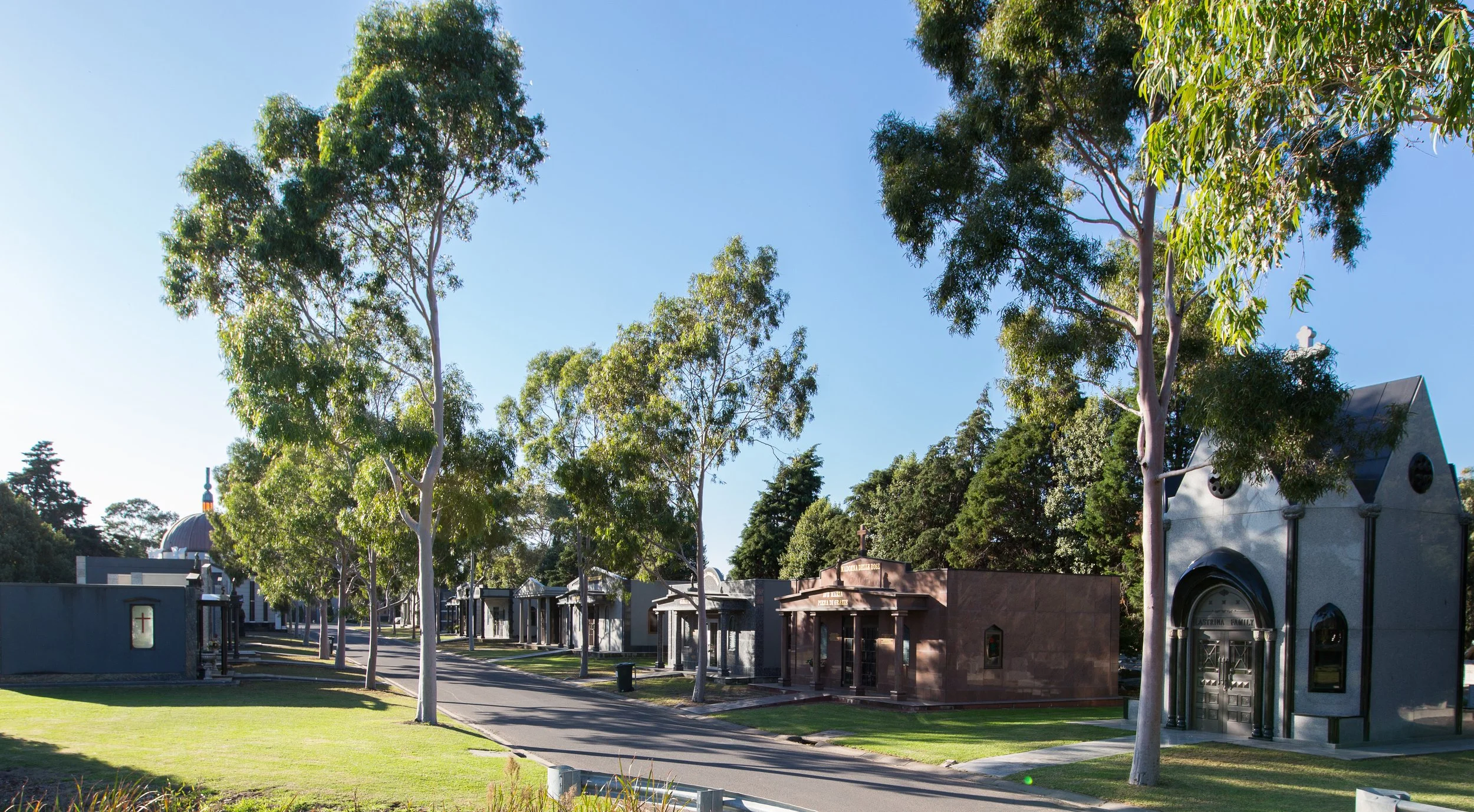 A cemetery with multiple small mausoleums and tombs, lined along a paved walkway, surrounded by tall green trees under a clear blue sky.