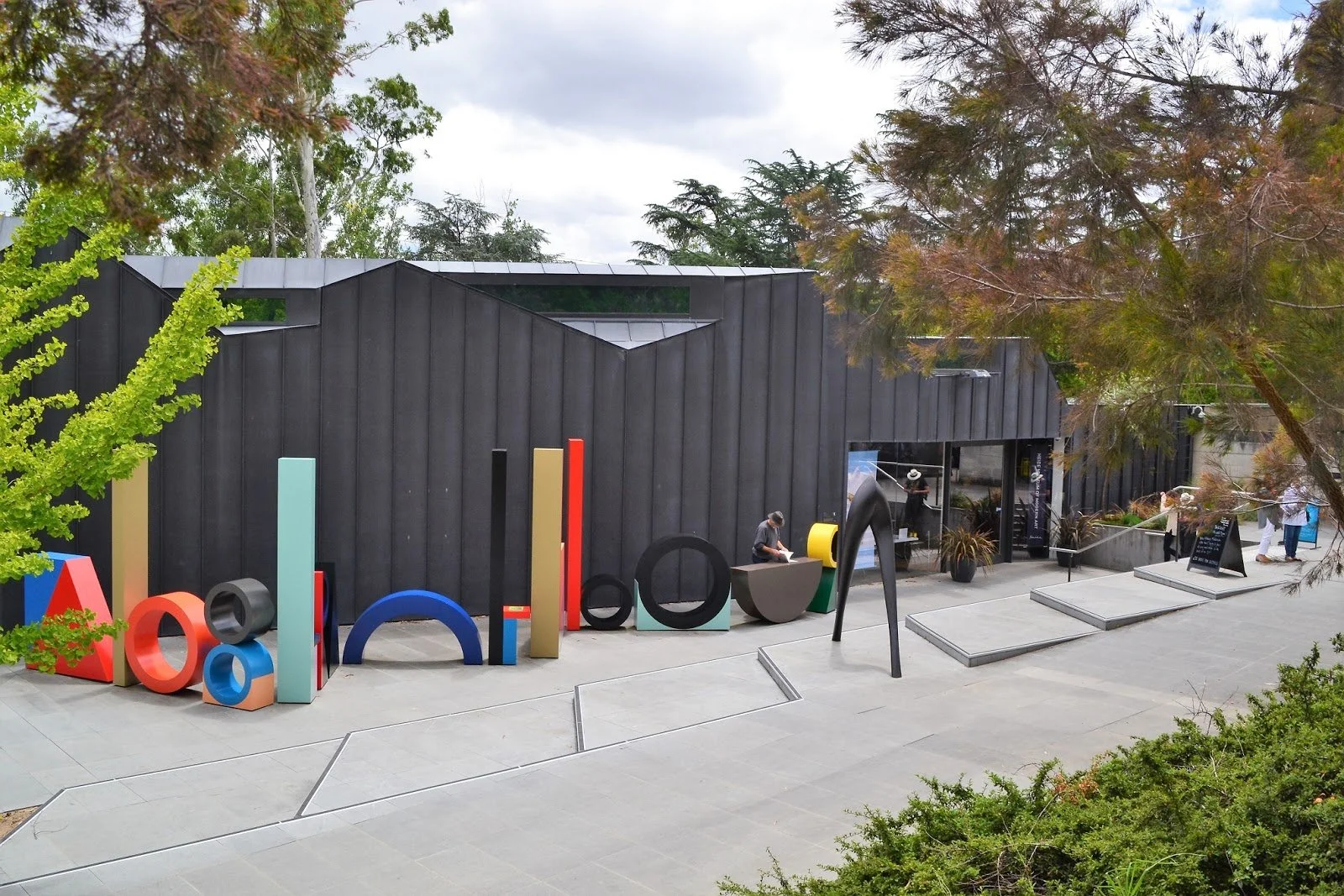 Exterior of an Art and Design Museum with colorful three-dimensional letter sculptures, a modern black building, trees, and people visiting the site.
