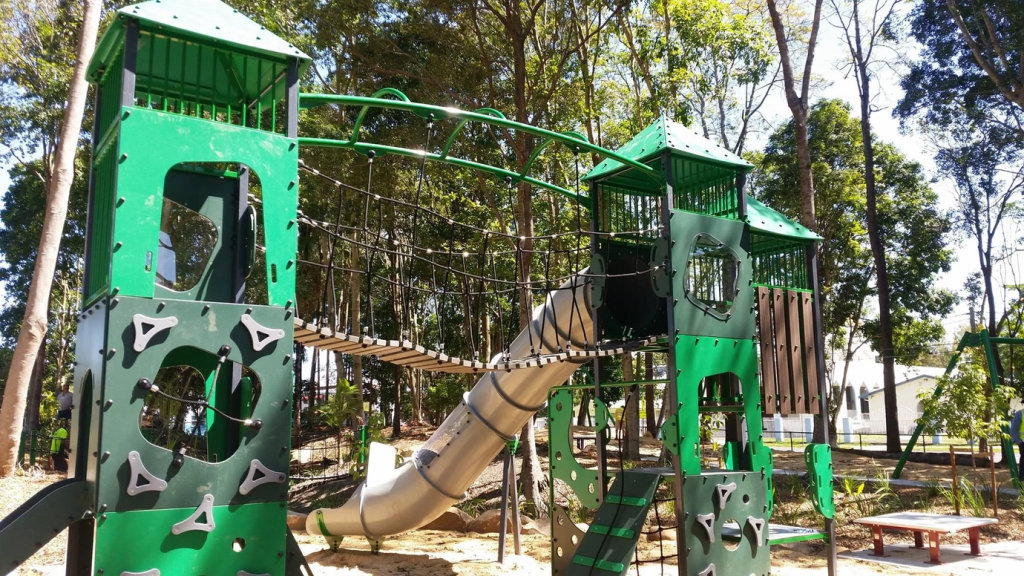 Colorful playground structure with slides, climbing areas, and bridges in an outdoor park setting surrounded by trees.