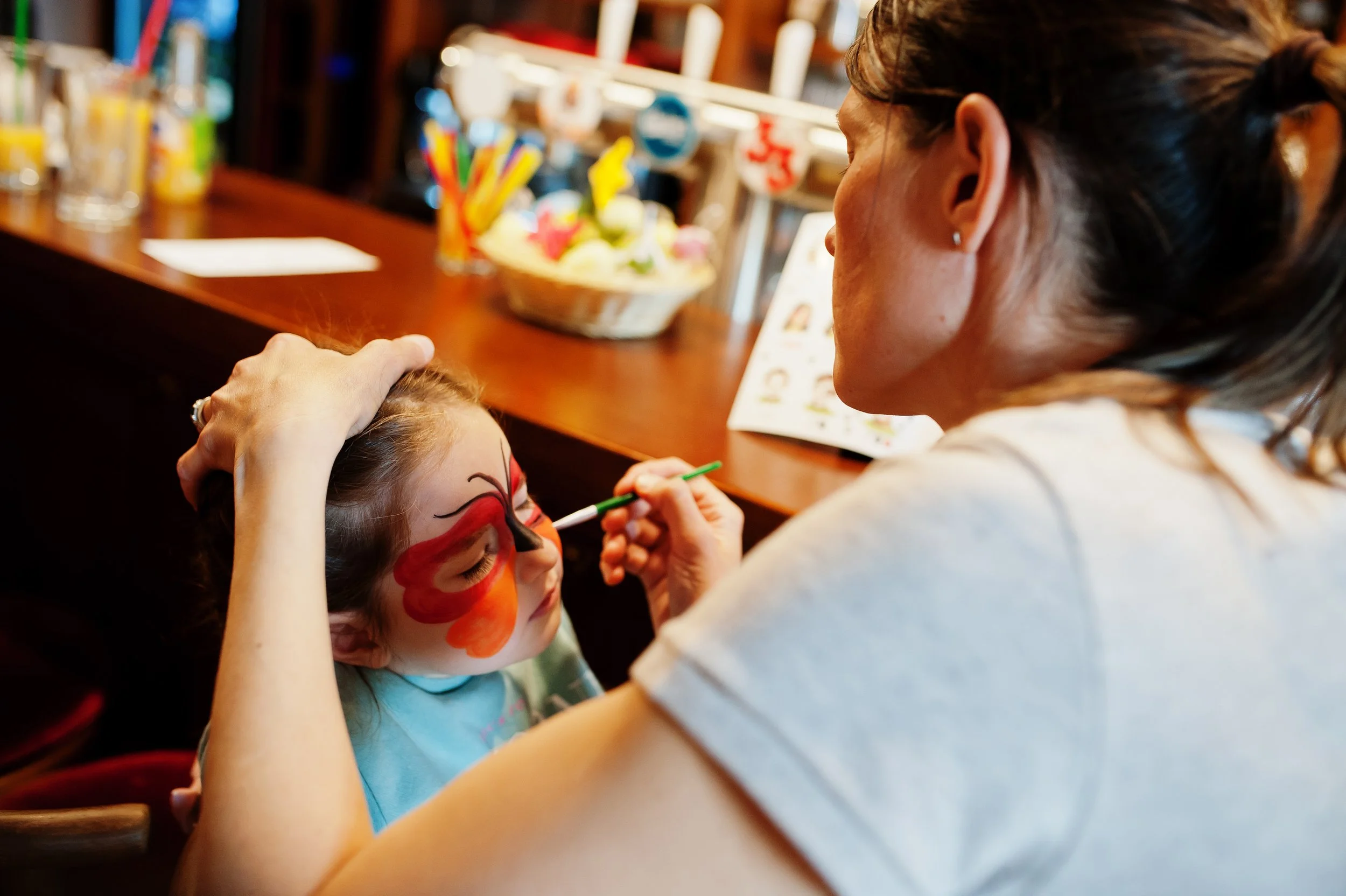 A woman is painting a colorful butterfly face paint on a young girl with her eyes closed, indoors at a table with face paint supplies in the background.