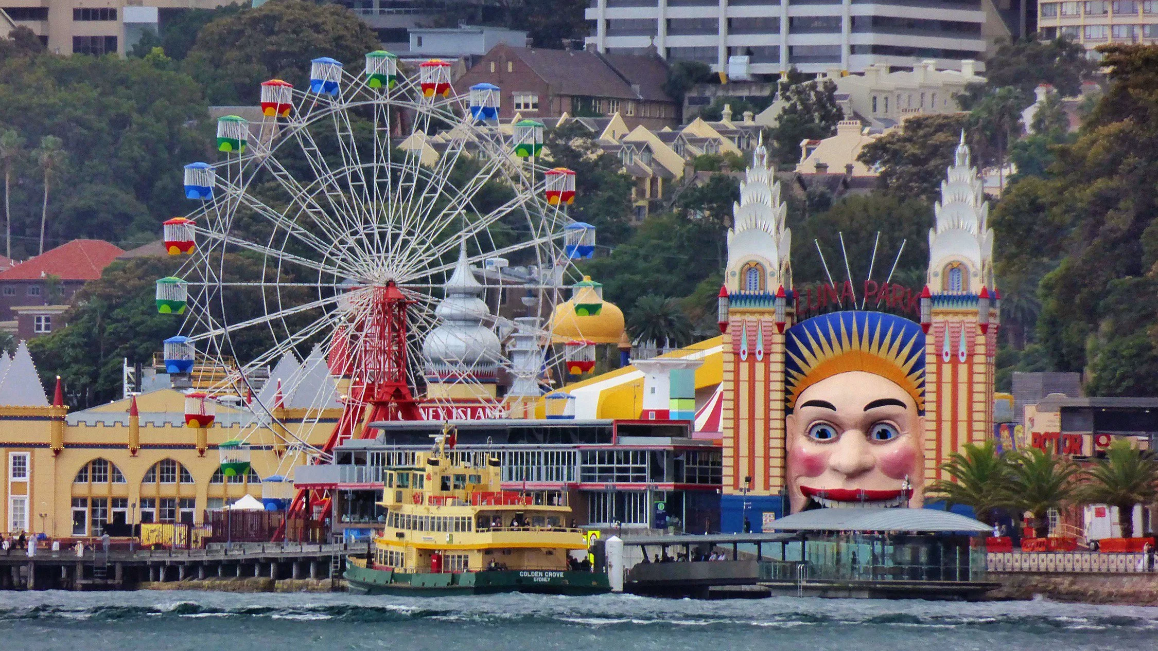 A colorful amusement park with a large ferris wheel, a giant face structure with a smiling expression, and surrounding rides and buildings along the waterfront with boats, set against a hillside with residential buildings and trees.