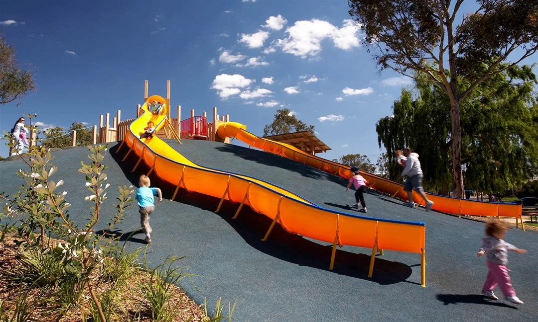 Children playing on a colorful outdoor slide in a park during daytime with a blue sky and trees in the background.