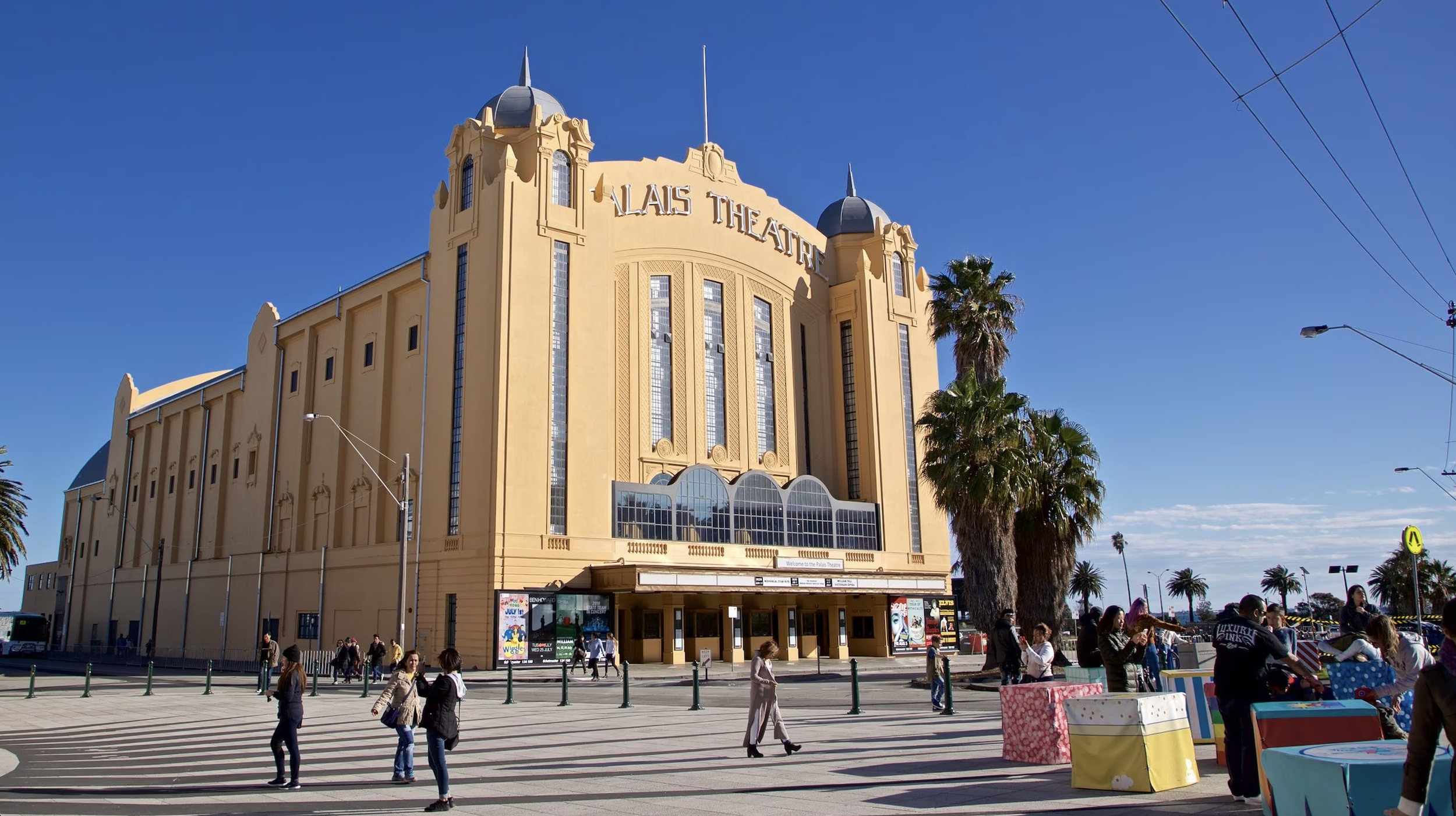 The historic Palace Theatre with a beige facade, tall vertical windows, and rounded towers topped with pointed spires, surrounded by palm trees and people walking and shopping outside on a sunny day.