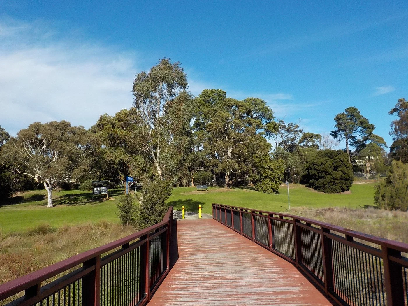 A wooden bridge with black metal railings spanning over a grassy wetland, leading to a park with trees and a clear blue sky.