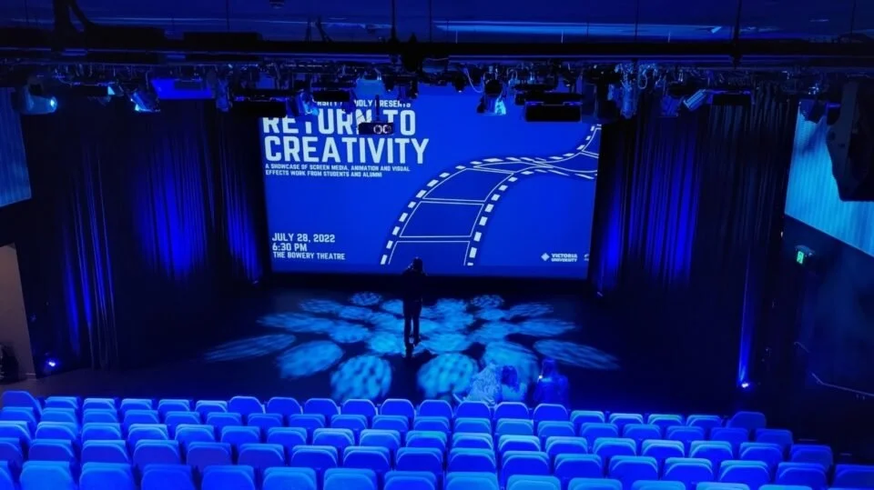 Empty theater with blue lighting, a large screen displaying an event titled 'Return to Creativity' scheduled for July 28, 2022, at 6:30 PM at The Bowery Theatre, with a person standing on stage.