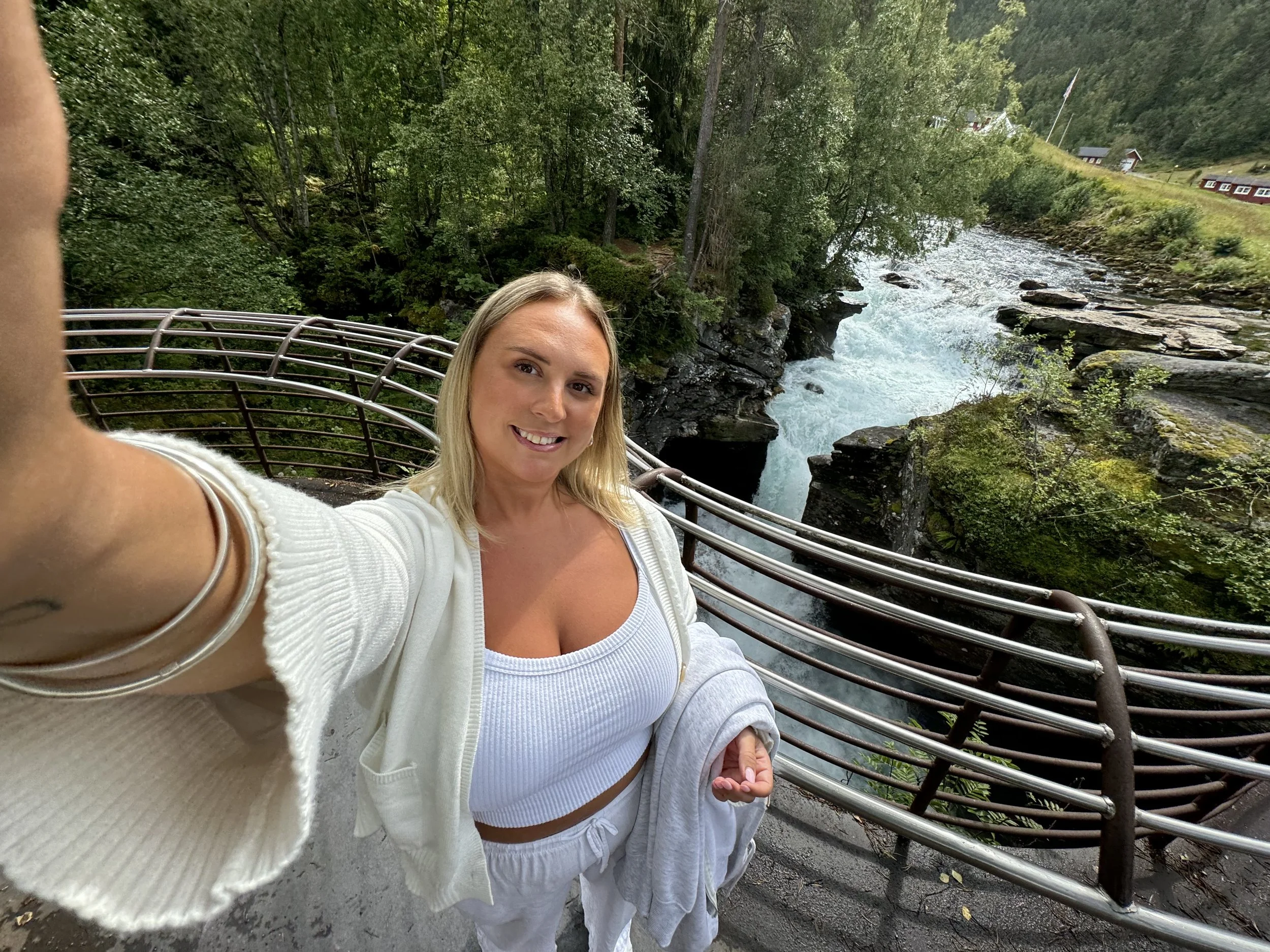 A woman taking a selfie on a bridge overlooking a river with rocks and green trees in a lush outdoor setting.