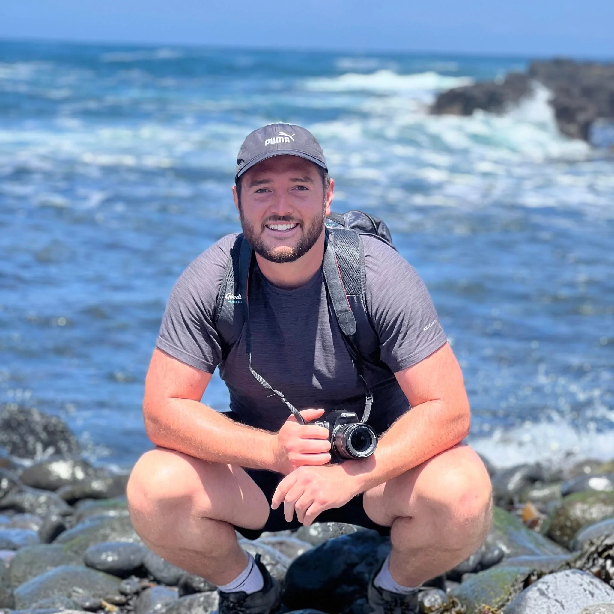 A man in a gray t-shirt and black shorts crouches on rocks by the ocean, holding a camera, smiling at the camera with a blue sky and waves in the background.