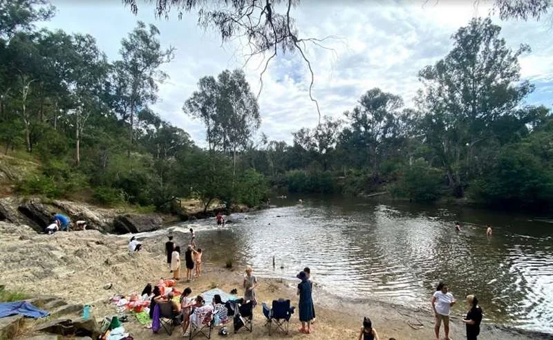 People relaxing and swimming at a riverbank surrounded by trees on a partly cloudy day.