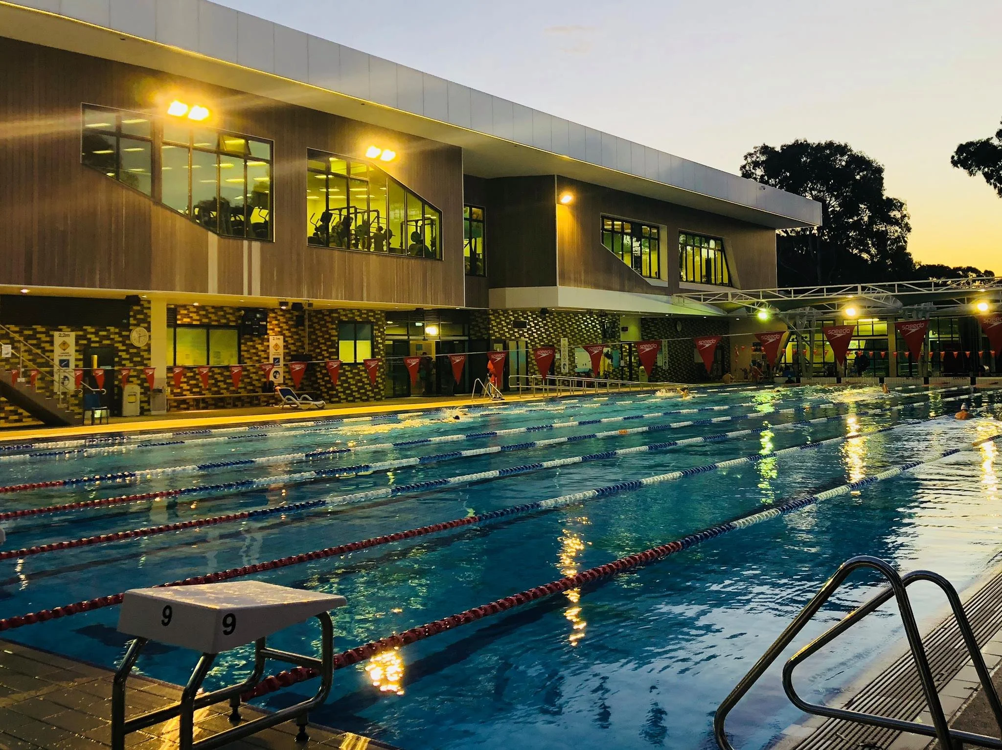An outdoor swimming pool area at dusk with people swimming, a two-story building with large windows showing gym equipment inside, and trees in the background.