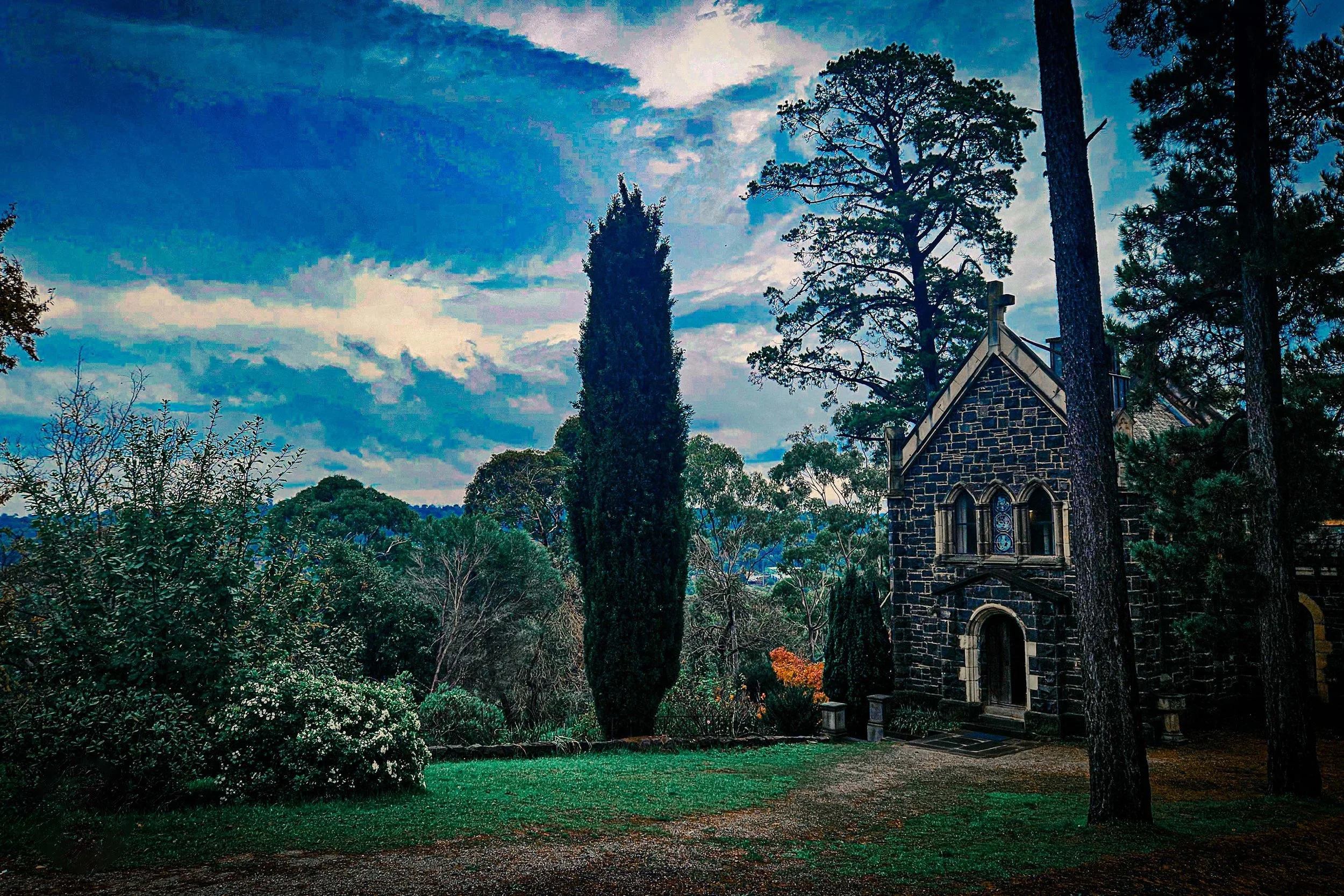 A small dark stone church surrounded by lush green trees and a well-kept lawn, under a cloudy sky at dusk.