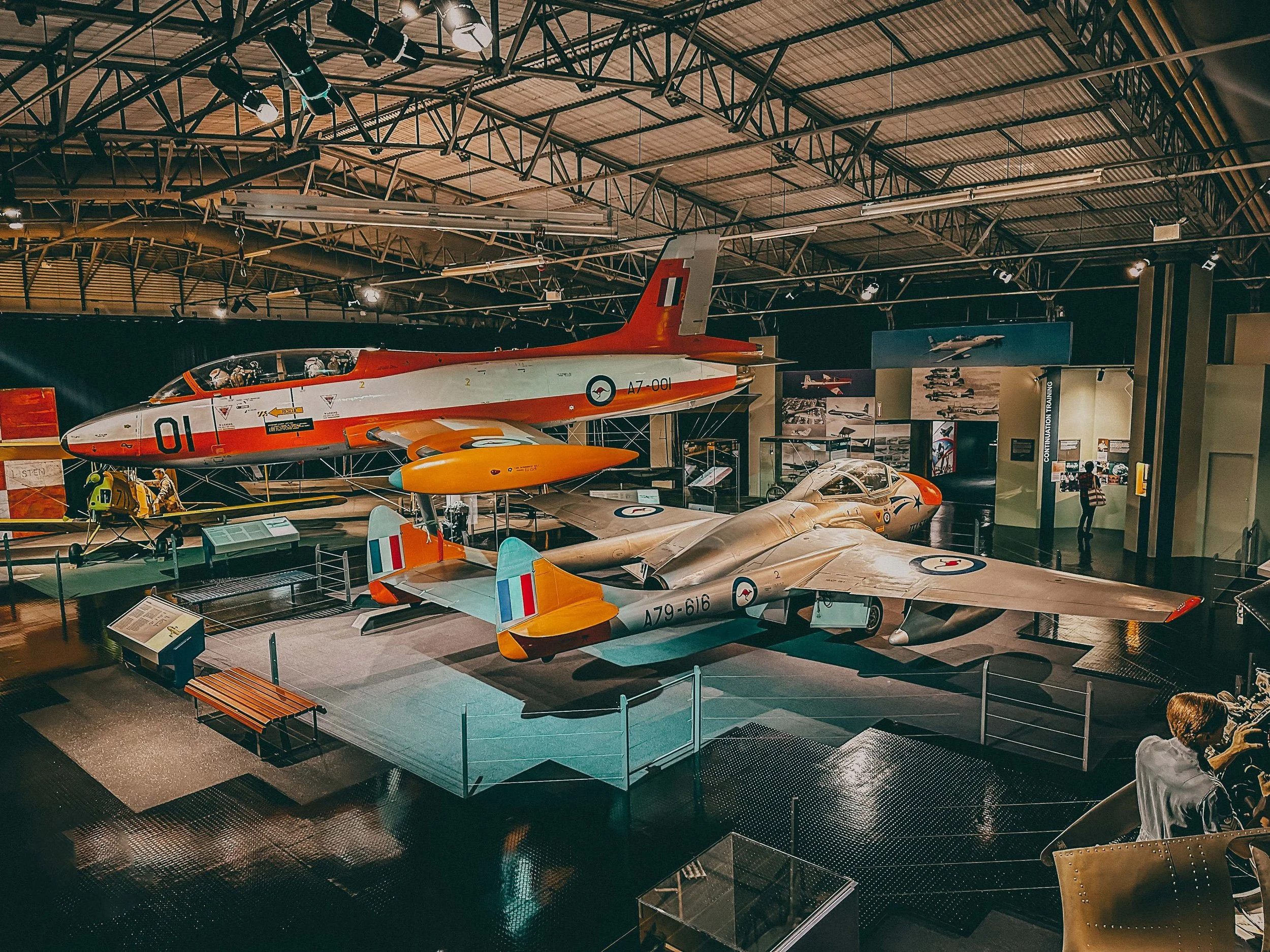 Two vintage fighter jets displayed in a museum, one on the floor with a sleek silver body and the other suspended overhead with a red and white body.