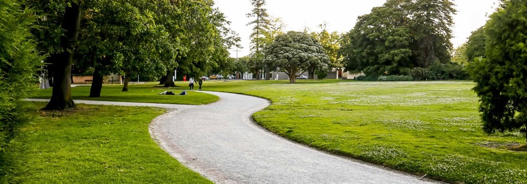 A winding gravel path through a lush green park with trees and a few people walking in the distance.