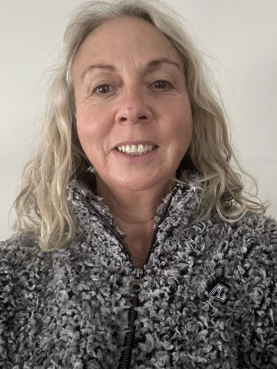 A woman with wavy blonde hair smiling at the camera, wearing a gray, fluffy zip-up jacket with a small logo on the chest, standing against a plain white background.
