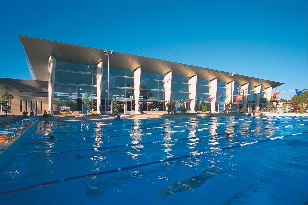 An outdoor swimming pool in front of a modern building with large glass windows, under a clear blue sky.