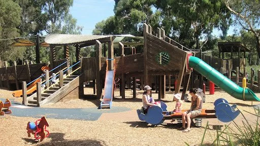 Children playing on a playground with slides, climbing structures, and seesaws, surrounded by trees and shaded areas.