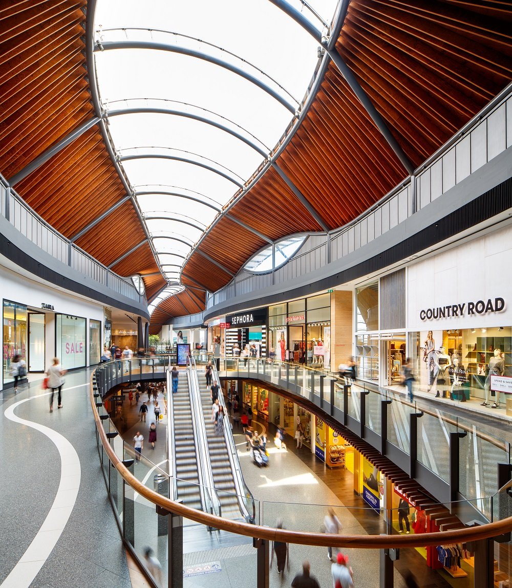 Interior view of a modern shopping mall with a curved wooden ceiling, escalators, and retail stores including Zara, Sephora, and Country Road. People are walking and shopping on both the upper and lower levels.