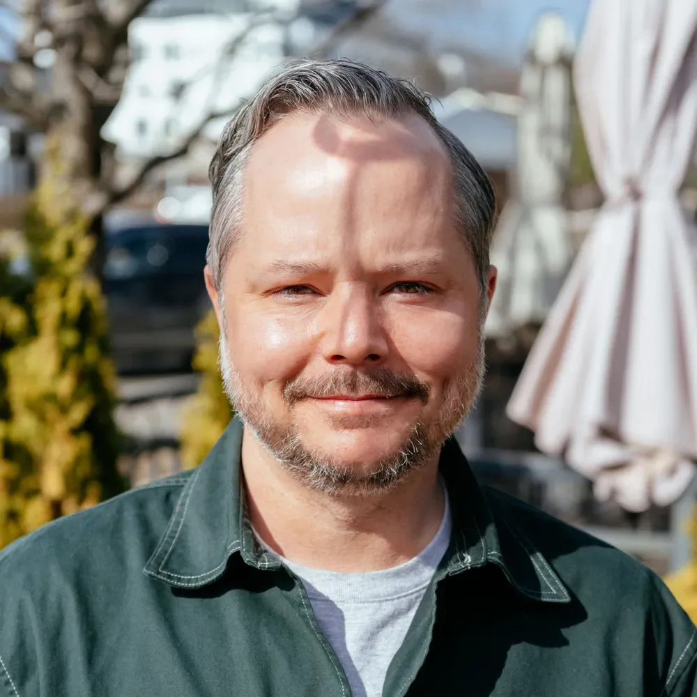 A middle-aged man with a beard smiling outdoors on a sunny day, with a blurred background of trees, buildings, and patio umbrellas.