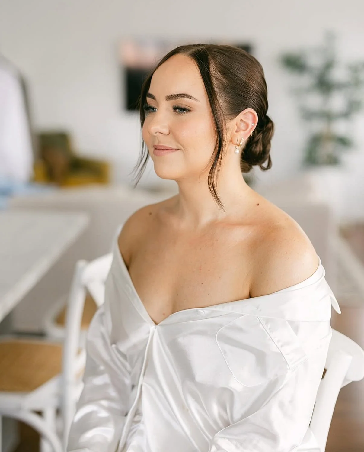 A woman with brown hair styled in a low bun, wearing earrings and a white satin off-shoulder top, sitting in a room with a blurred background.