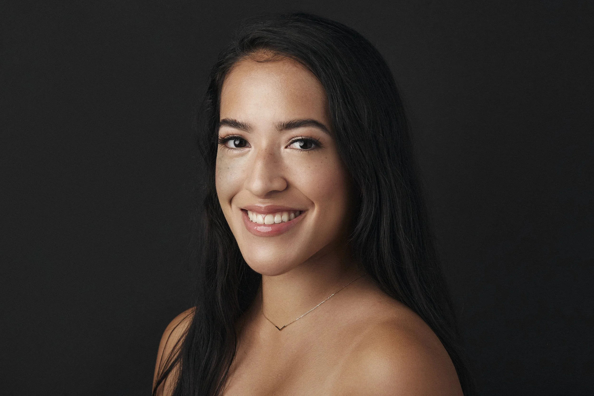 Close-up portrait of a young woman with long dark hair, smiling, wearing a delicate necklace, against a dark background.