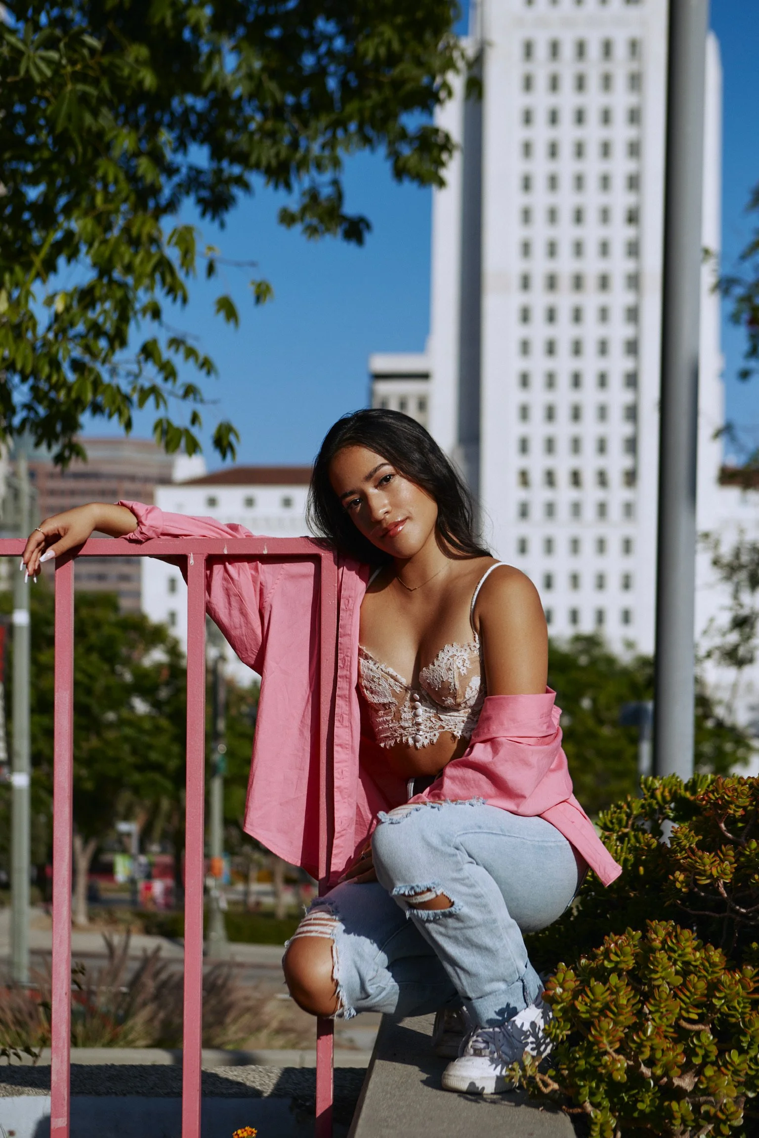 A young woman with long dark hair crouches next to a pink railing outdoors, wearing a pink jacket, a lace top, ripped jeans, and white sneakers, against a backdrop of tall white buildings and green trees on a sunny day.