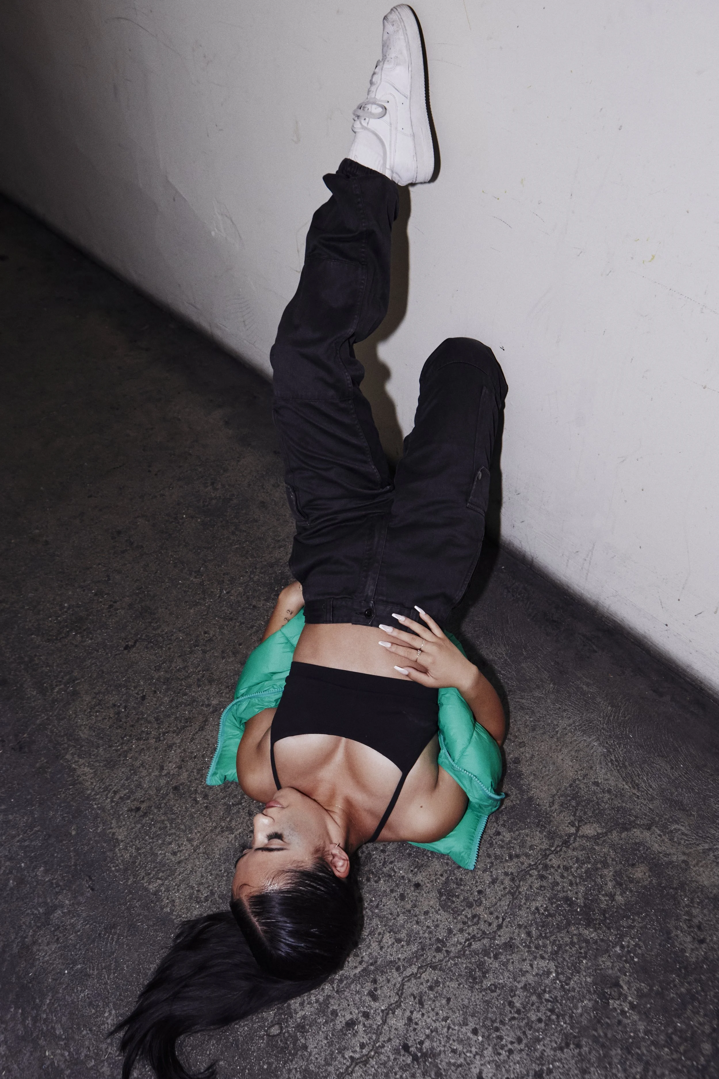 A woman lying on the ground with her head near the floor and her legs against the wall, wearing a black tank top, black pants, a green jacket, and white sneakers.