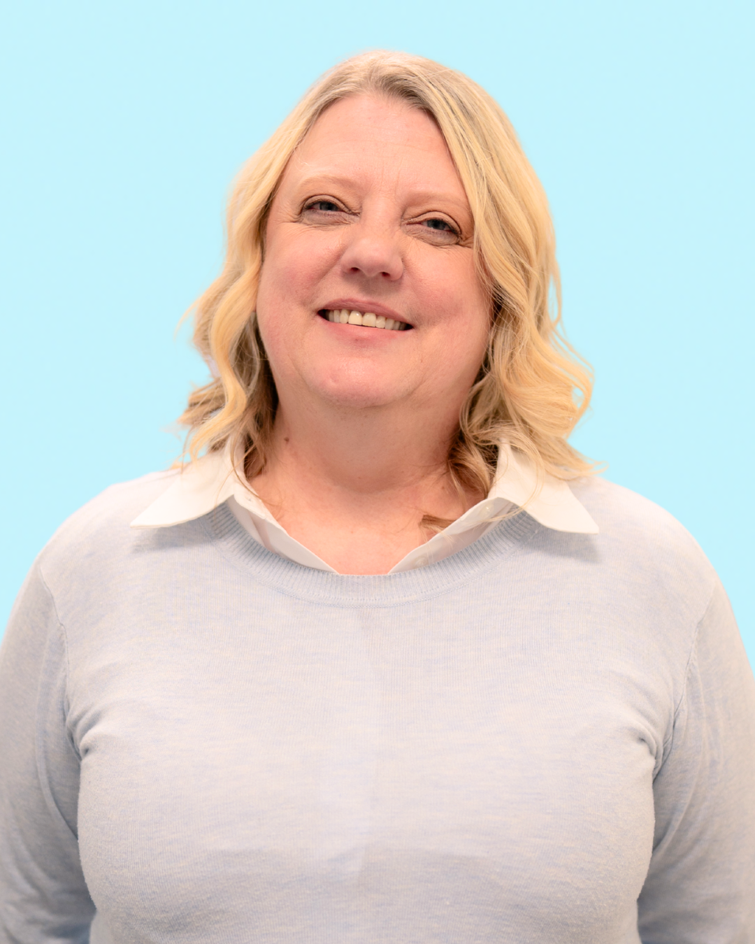 A woman with blonde, curly hair smiling, wearing a white collared shirt under a light gray sweater, against a light blue background.