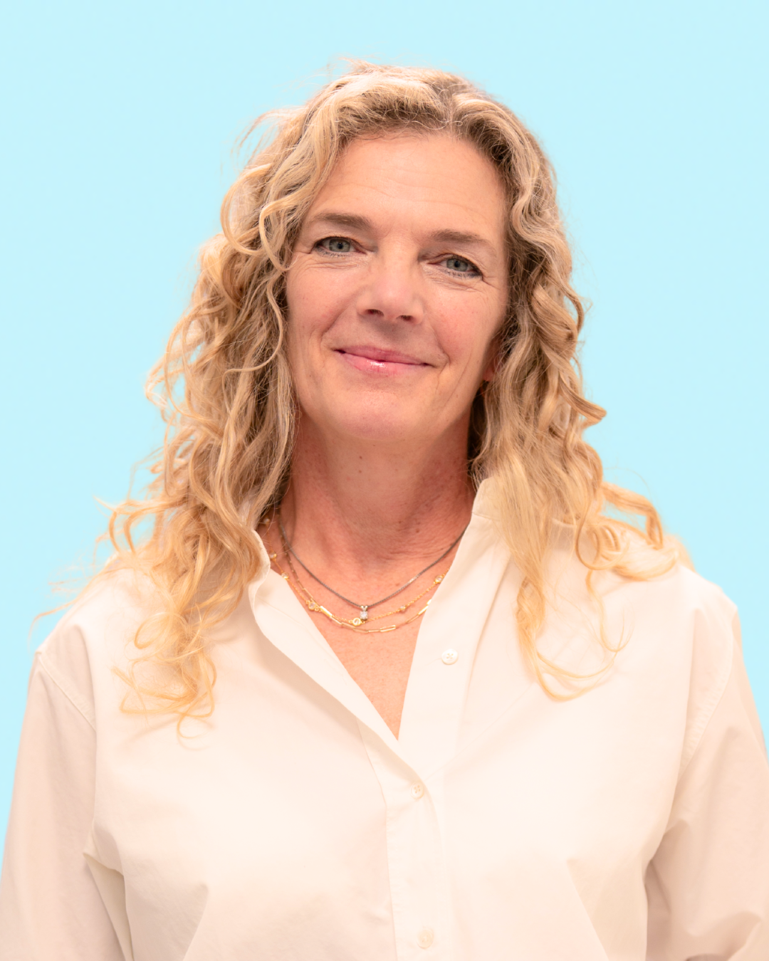 A woman with blonde curly hair smiling, wearing a white button-up shirt and layered necklaces, against a light blue background.