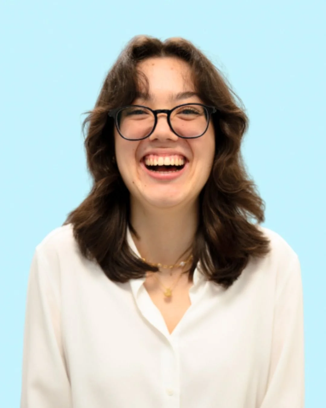 A young woman with glasses and shoulder-length brown hair, smiling widely, wearing a white blouse and a gold necklace, against a light blue background.