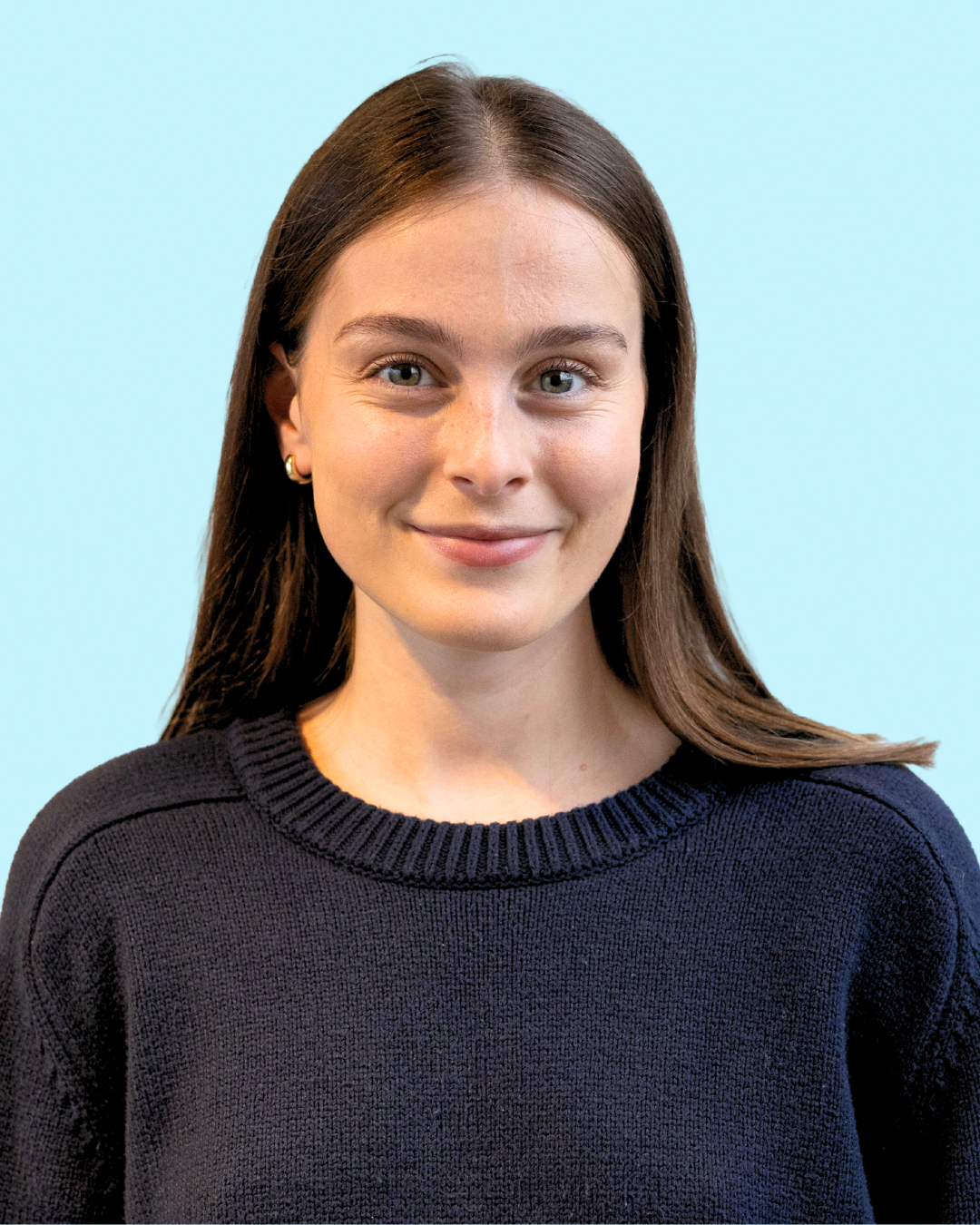 Portrait of a young woman with long brown hair, light skin, wearing a black sweater, smiling, against a light blue background.