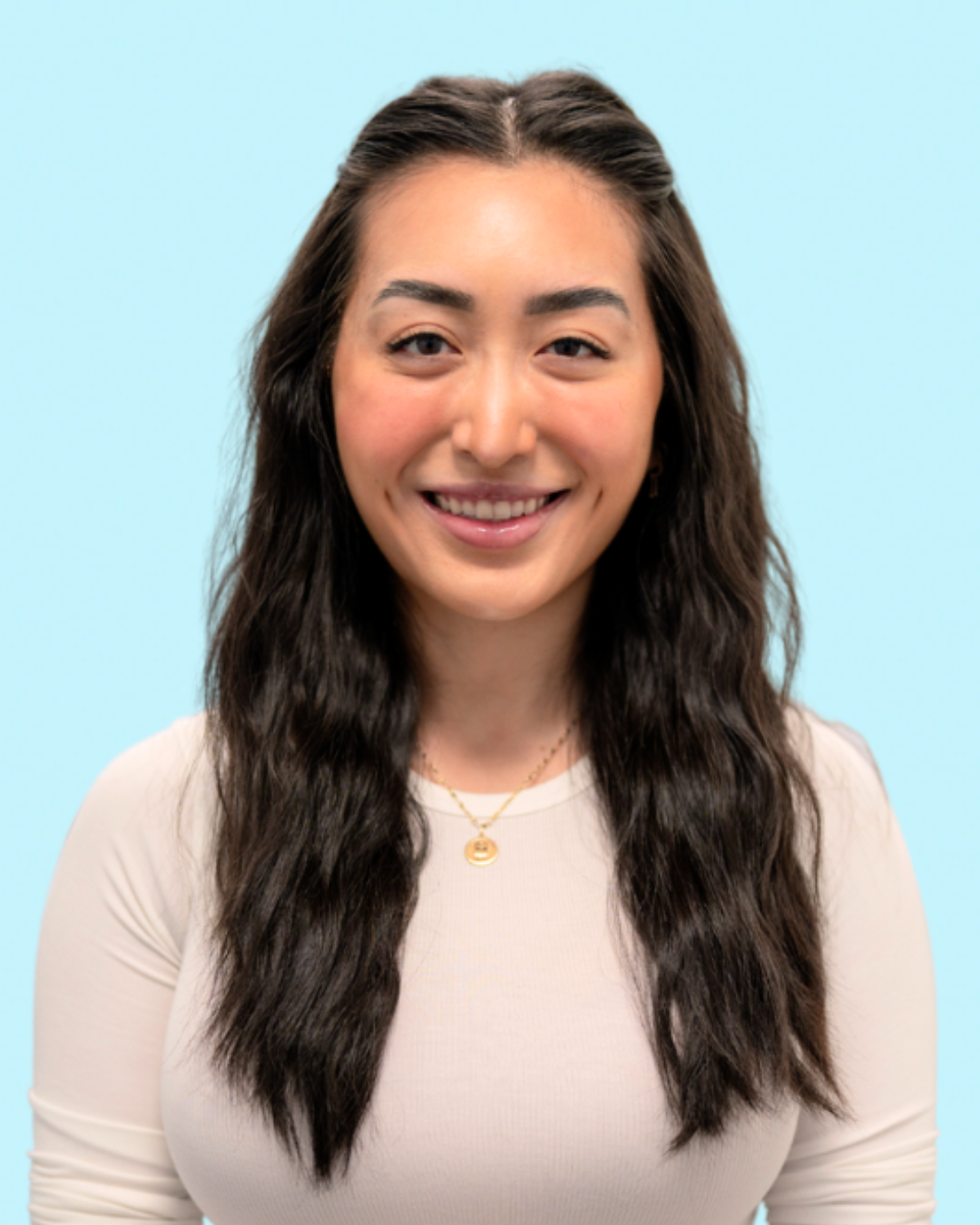 Portrait of a young woman with long, wavy dark hair, smiling, wearing a beige top and gold jewelry, against a light blue background.