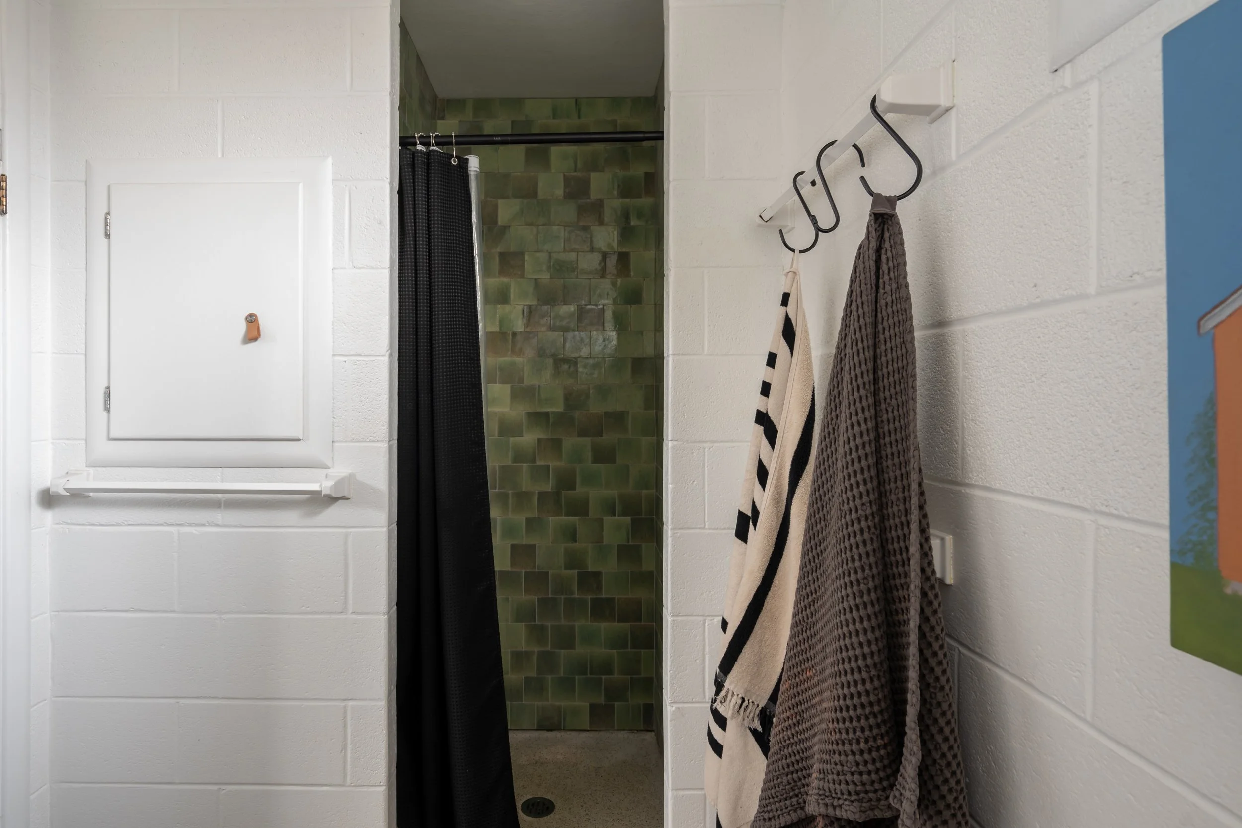 Inside a shower room with white brick walls and a green tiled shower area, black shower curtain, and towels hanging on hooks on the right wall.