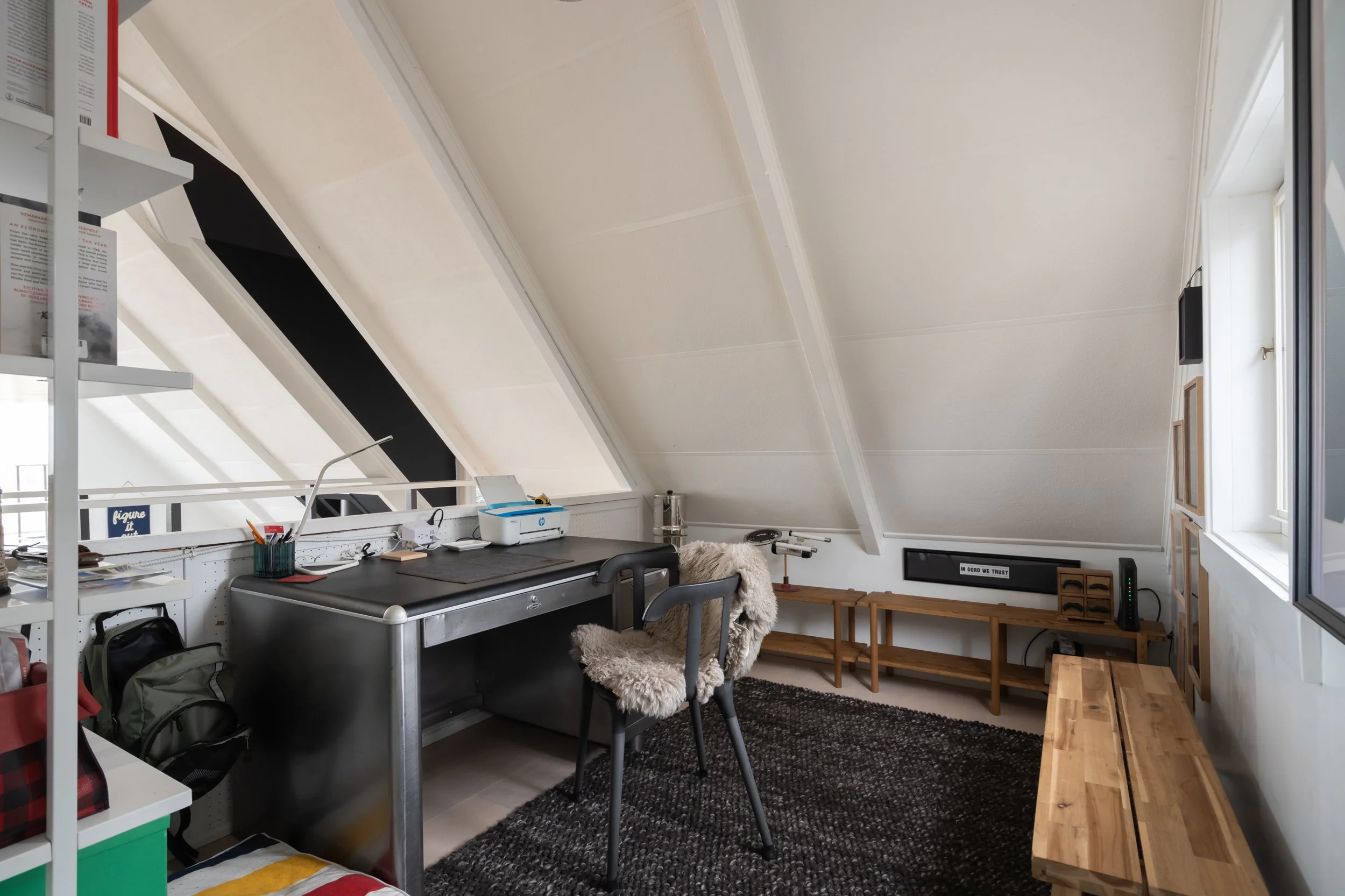 A cozy attic workspace with a sloped ceiling, a metal desk, a black chair with a furry throw, a wooden bench, shelves, and a window letting in natural light.