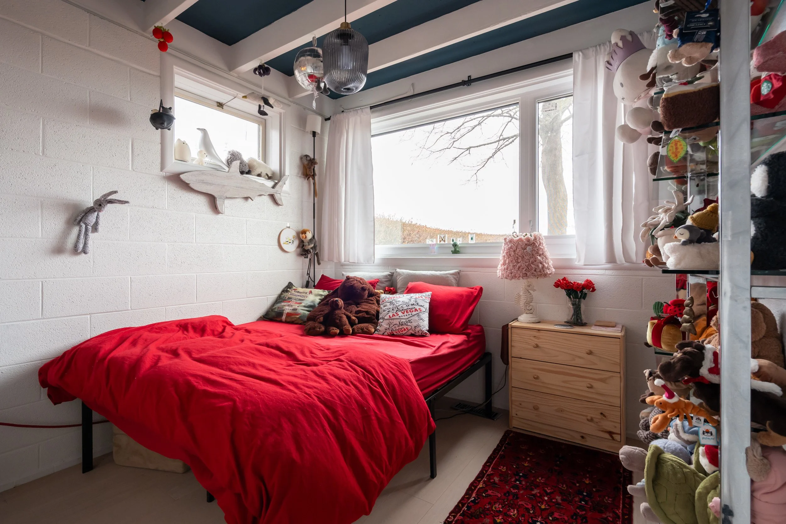 A cozy bedroom with a white painted brick wall, a window with white curtains, and a bed with red bedding. The bed has plush toys, including a teddy bear. To the right, there's a wooden nightstand with a pink flower lamp and a vase of red flowers. On 