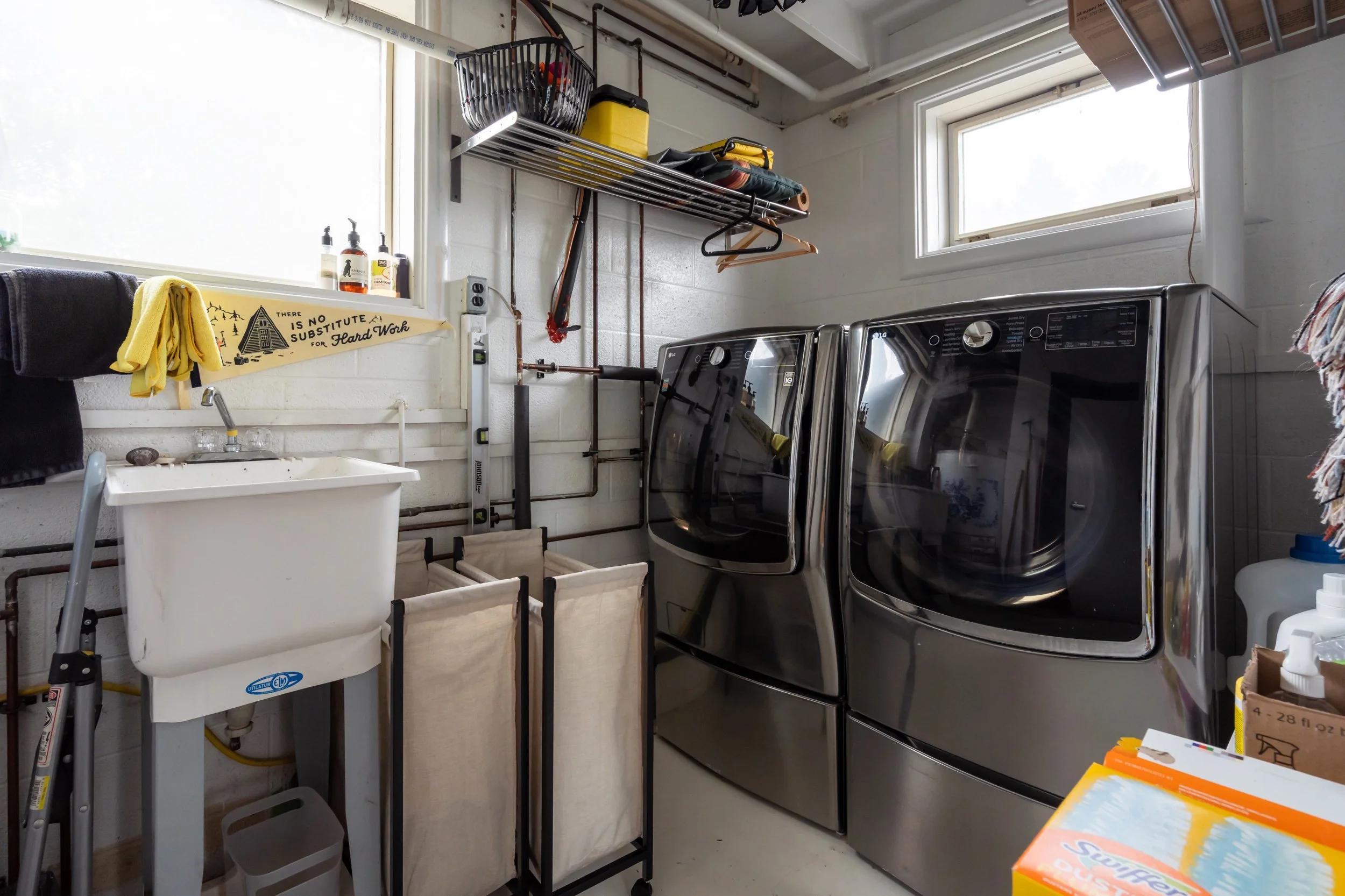 A laundry room with a white utility sink, a washing machine, and a dryer. There are shelves with cleaning supplies and storage bins, a window, and a humorous sign that says, "There is no substitute for hard work."