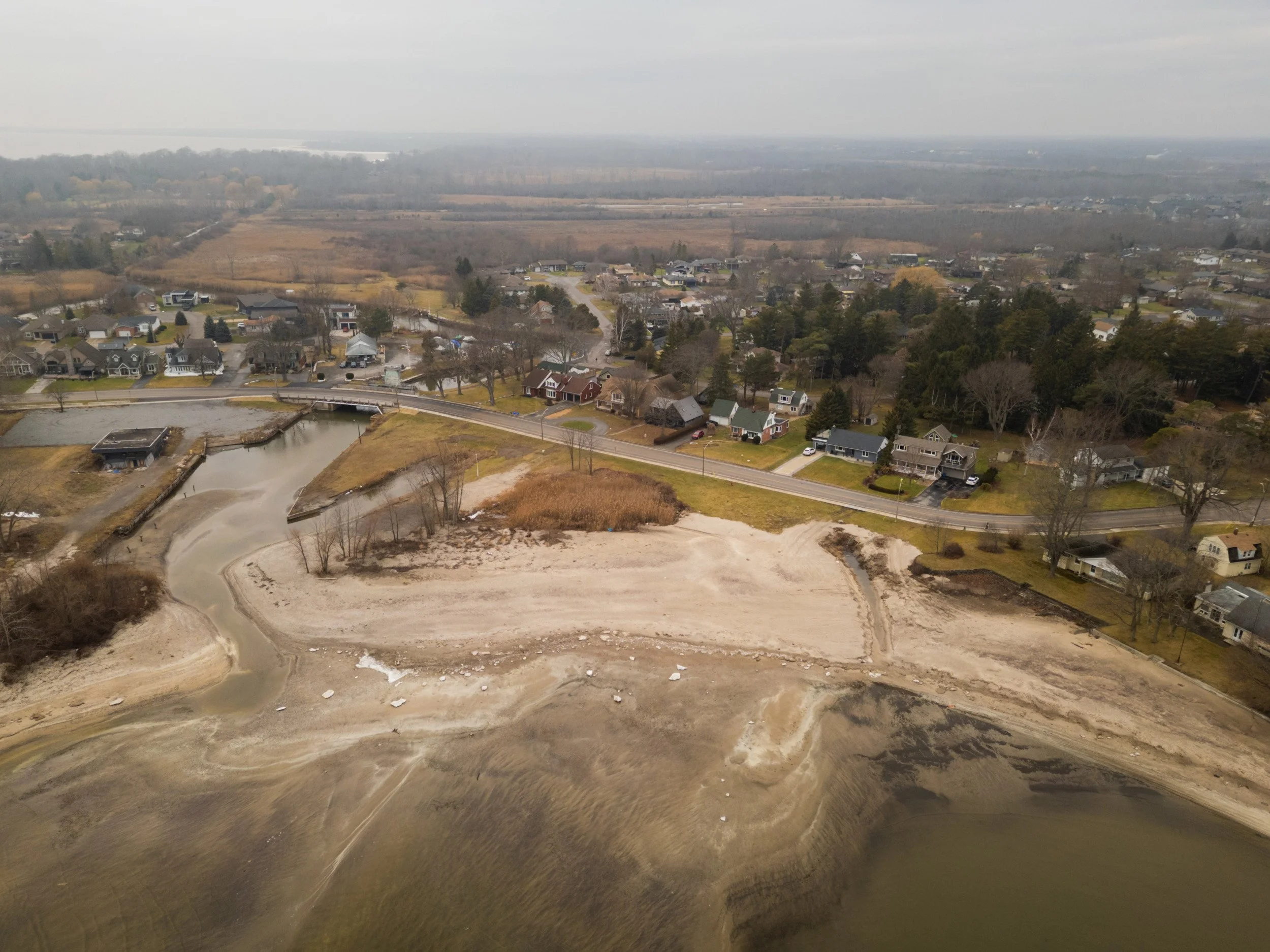 Aerial view of a residential neighborhood near a body of water with a sandy, barren area in the foreground, overcast sky.